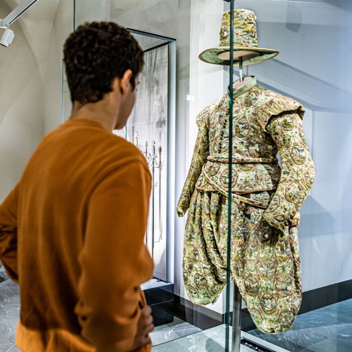 Junge schaut auf ein grünes, detailreich verziertes Gewand mit einem dazu passenden Hut in der Ausstellung Macht und Mode 
© SKD, Foto: Carina Sonntag